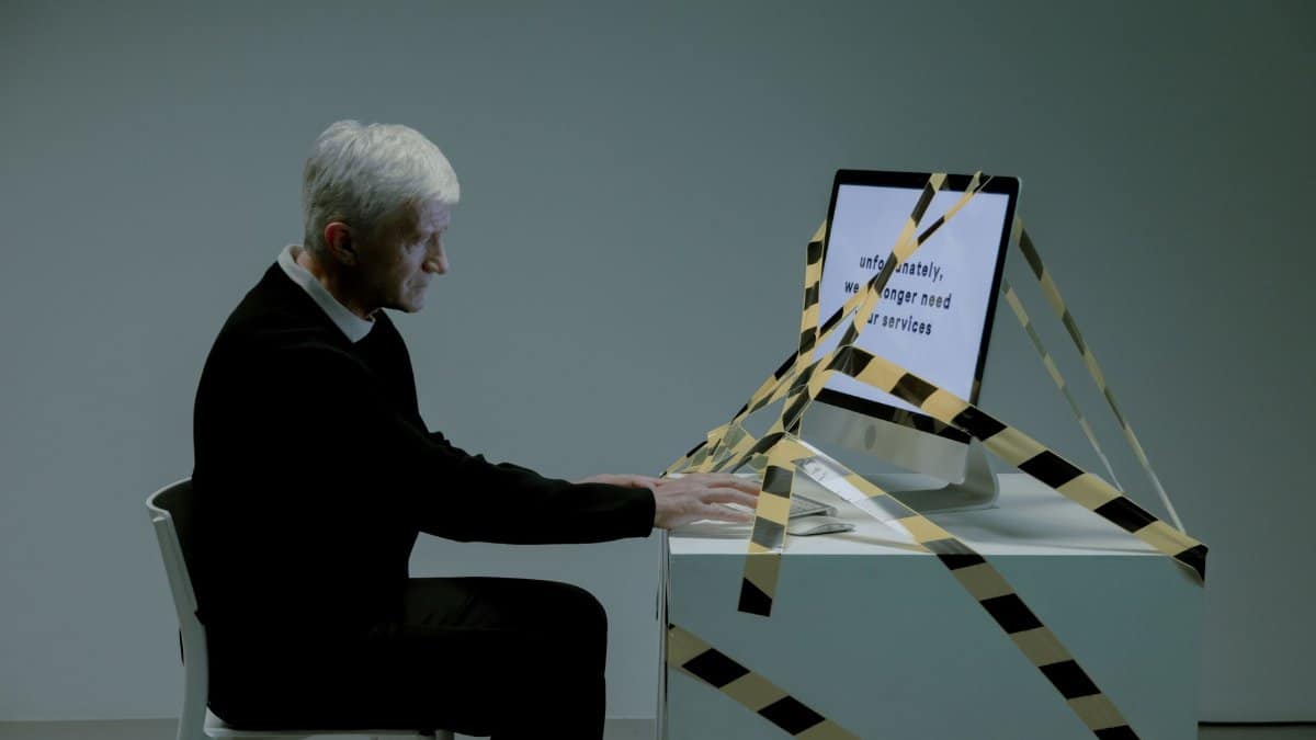 An elderly man sits at a desk with a computer, dealing with job termination notice.