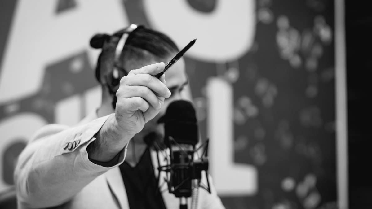 Man engaged in a studio broadcast holding a pen, in black and white.