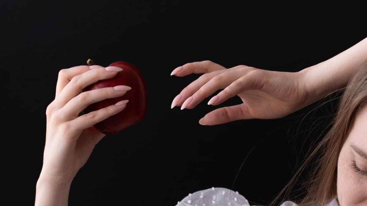 Elegant woman's hand holding a red apple against a dark background, symbolizing temptation.