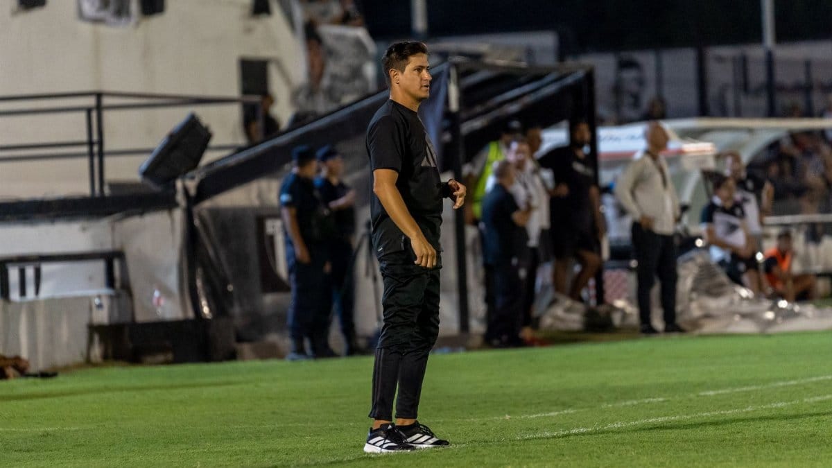 Focused football coach stands on the field sidelines during an intense match at night.