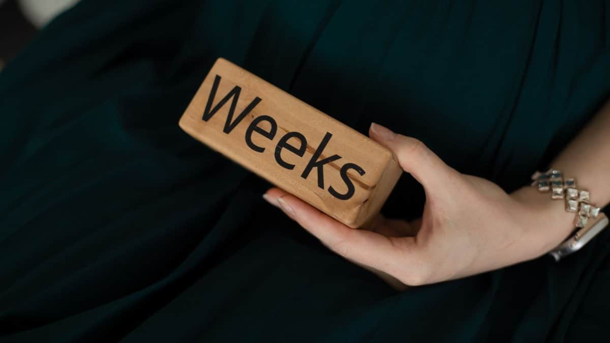 Close-up of a woman's hand holding a wooden block with the text 'Weeks,' on a green background.