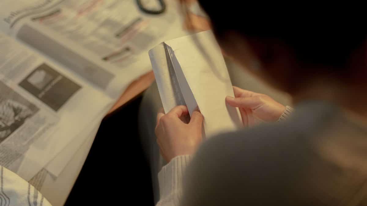 A woman reviews job postings with an envelope and newspaper inside a warm-lit room.