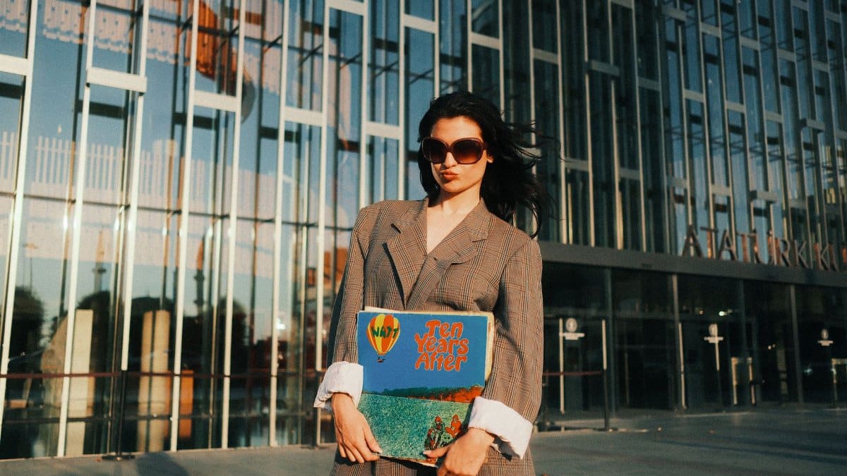 Fashionable woman with sunglasses and vinyl album standing outside a modern glass building at sunset.
