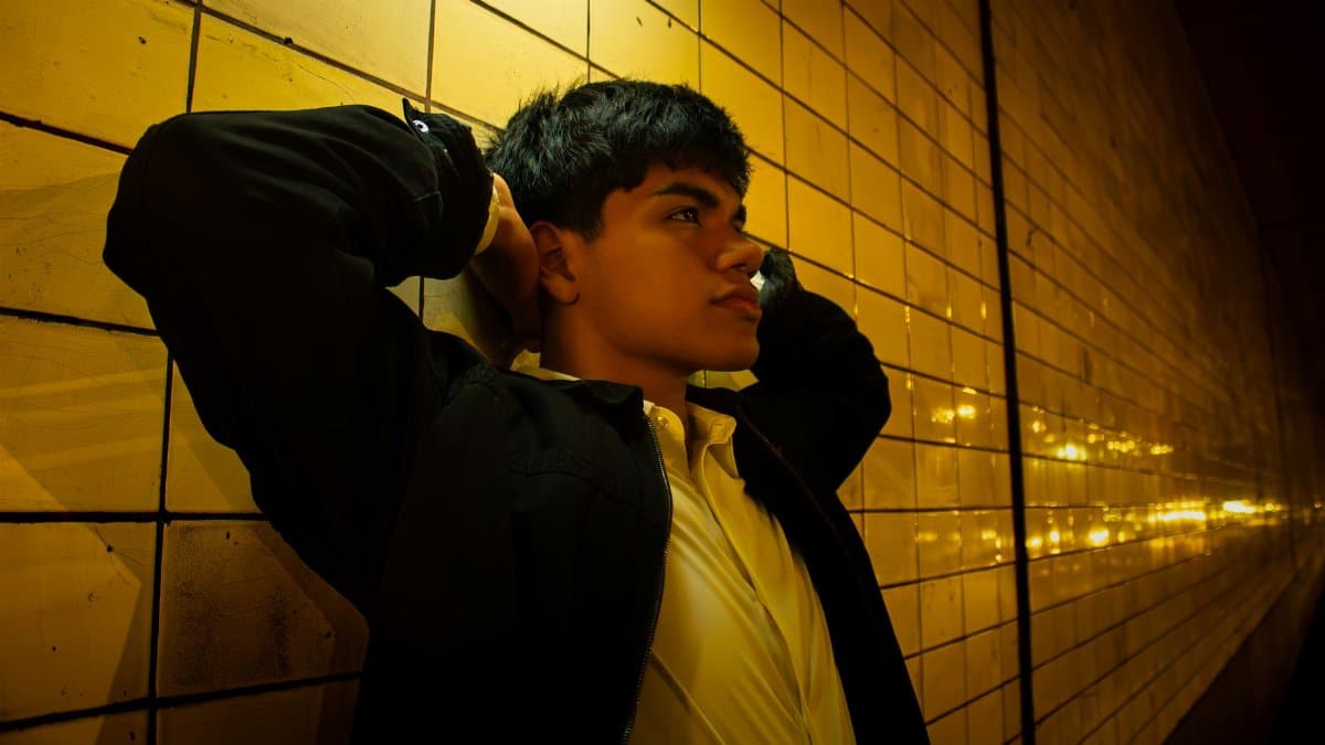 A young man in deep thought leaning against a warmly lit yellow tiled wall at night.