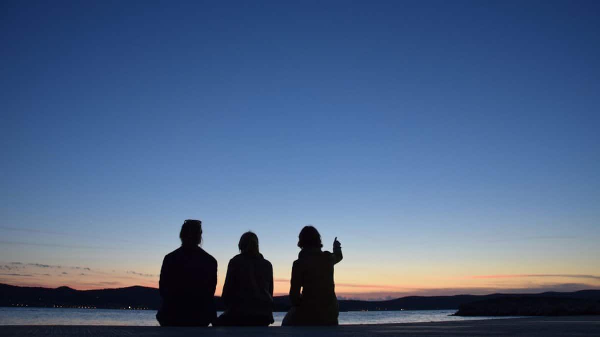 Three people sitting by a lake, enjoying a tranquil sunset view, creating perfect silhouettes.