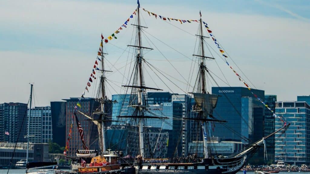 Historic USS Constitution sailing through Boston Harbor with city skyline in the background.