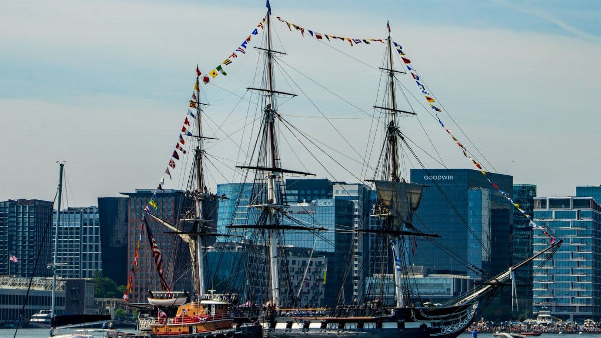 Historic USS Constitution sailing through Boston Harbor with city skyline in the background.