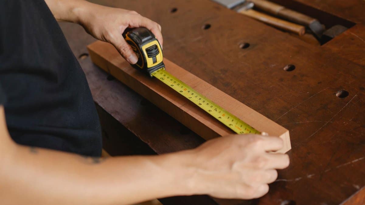 Precise unrecognizable male carpenter accurately measuring wooden detail while using ruler during work at workbench with instruments in professional studio
