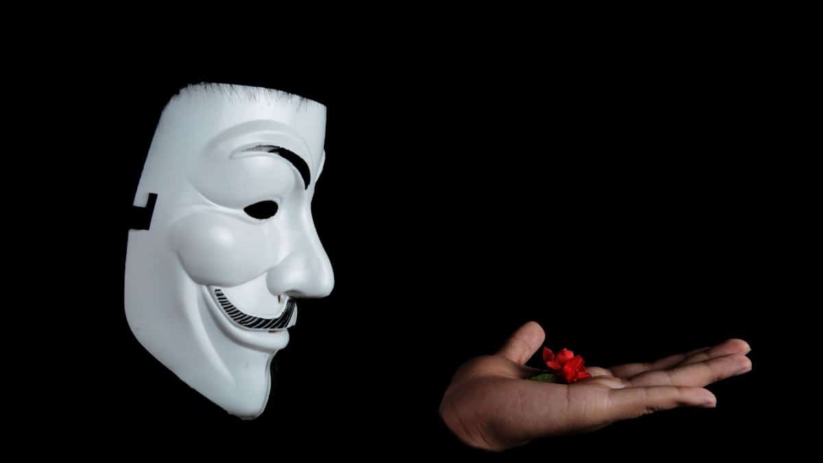 Side view of an anonymous mask facing a hand holding a red flower against a black background.