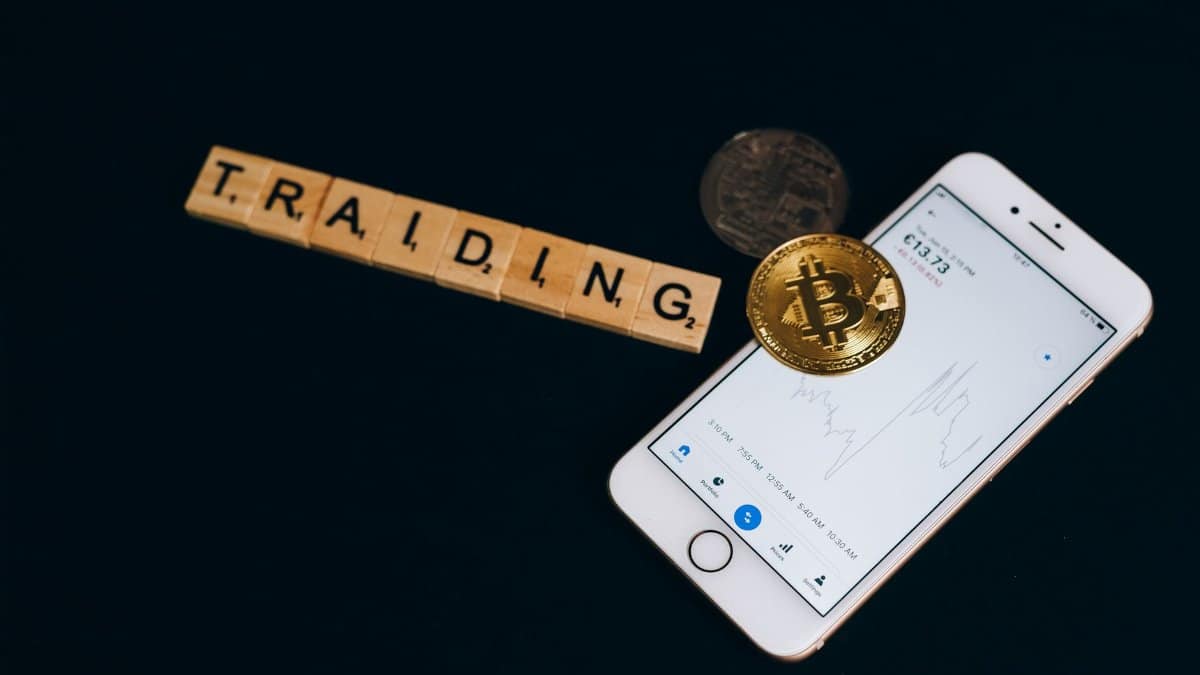 Cryptocurrency trading concept featuring Bitcoin, Scrabble tiles, and a smartphone on a black surface.