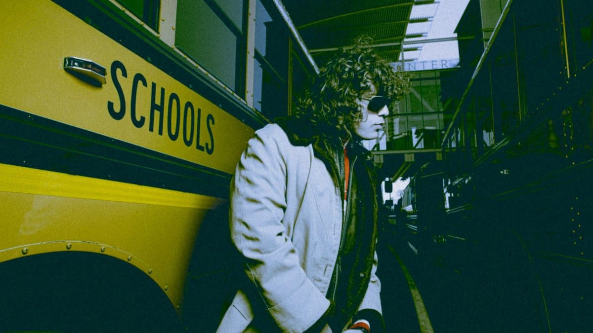 Man in coat and sunglasses standing next to a school bus in an urban setting.
