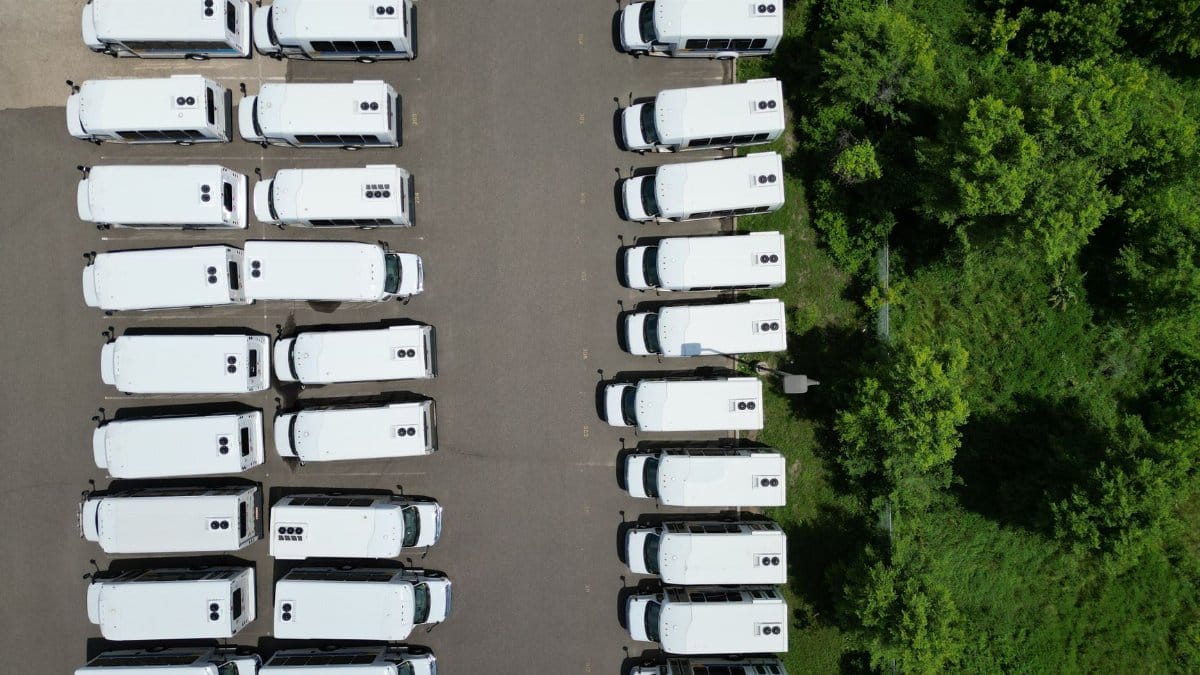 Aerial shot of a parking lot filled with white buses lined up next to a green forested area.