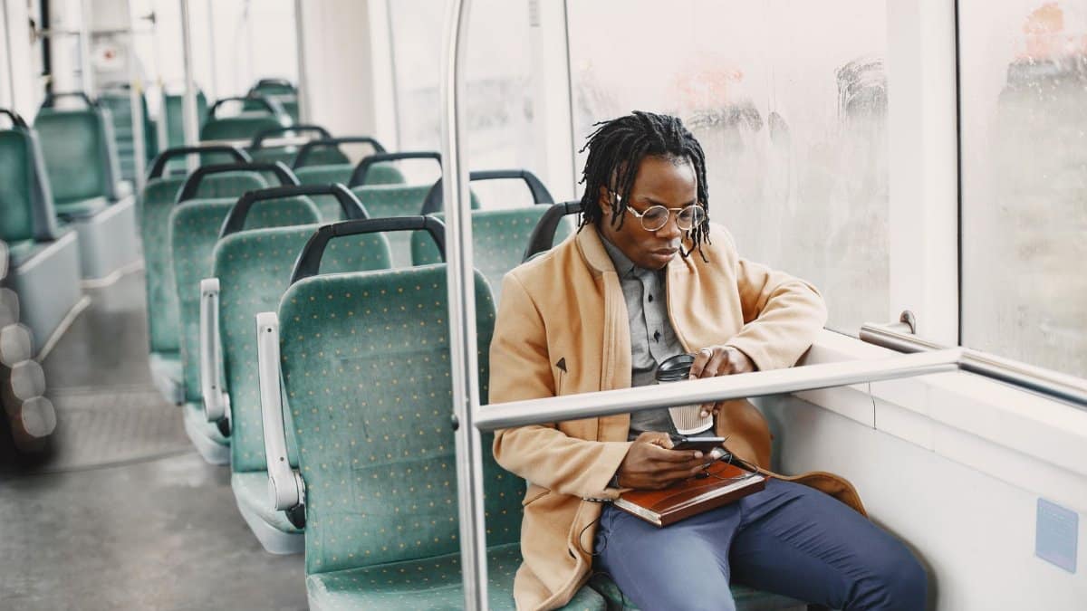 African American man on bus, wearing facemask and coat, using smartphone for commuting.