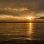 Dramatic golden hour sunset over calm ocean in Newport News, showcasing reflected tower silhouette.