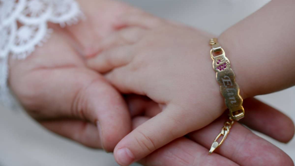 Close-up of a baby holding an adult's hand, featuring a gold bracelet with engraved name.