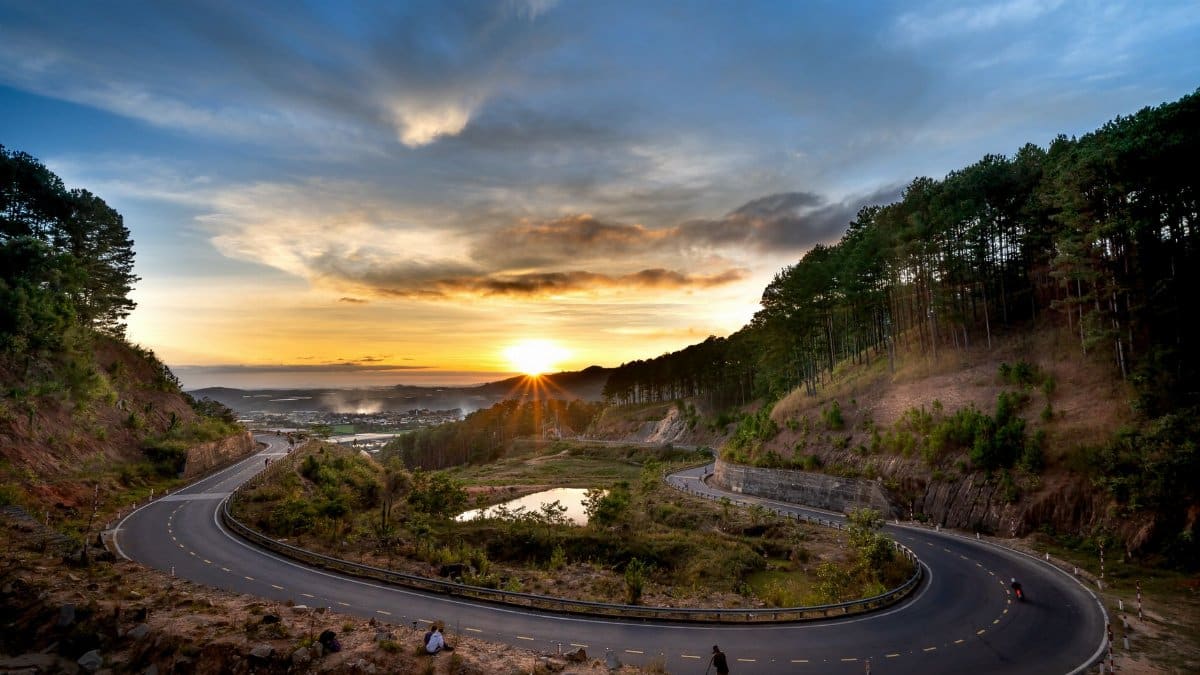 A winding road through the mountains at sunset, surrounded by lush forests and dramatic skies.