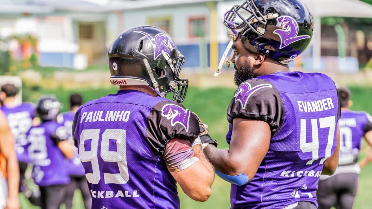 Two American football players in purple jerseys and helmets preparing on the field.
