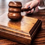 Close-up image of a wooden gavel held by hand on a desk in a courtroom setting.