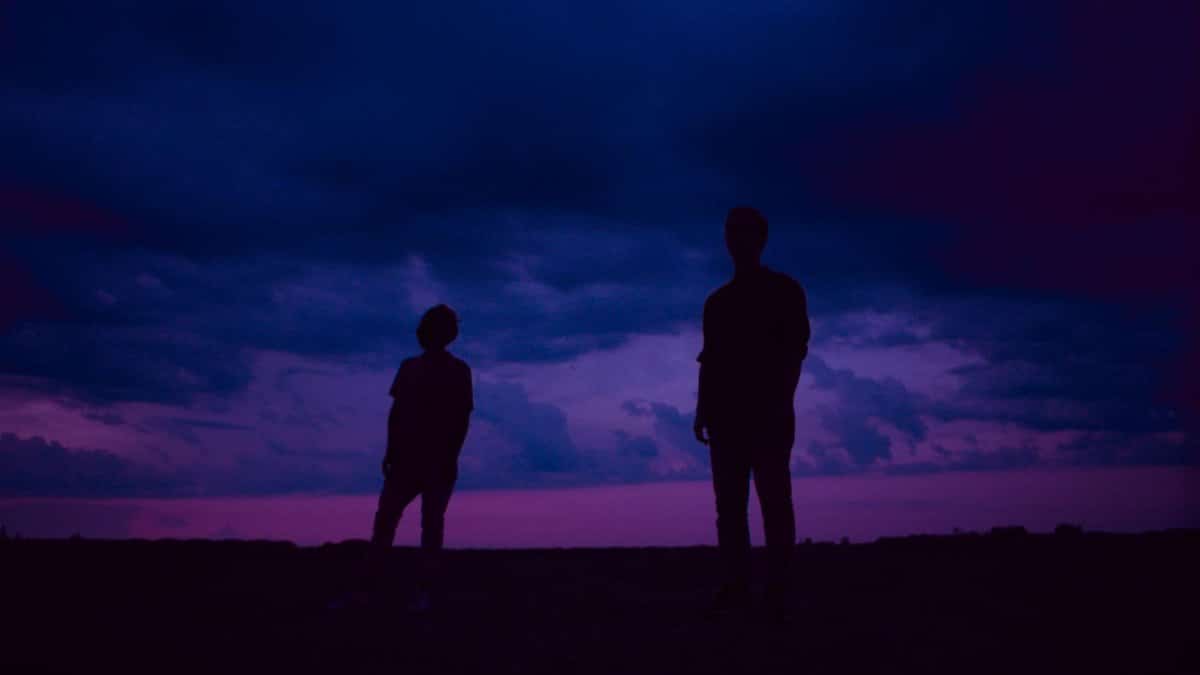 Two silhouettes stand beneath a dramatic twilight sky in a serene landscape.