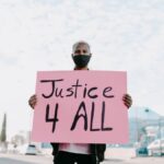Man holding a 'Justice 4 All' sign while wearing a face mask during an outdoor protest.