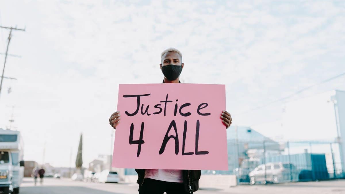 Man holding a 'Justice 4 All' sign while wearing a face mask during an outdoor protest.
