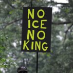 A protest sign with bold letters seen during a demonstration in Baltimore, surrounded by dense tree foliage.