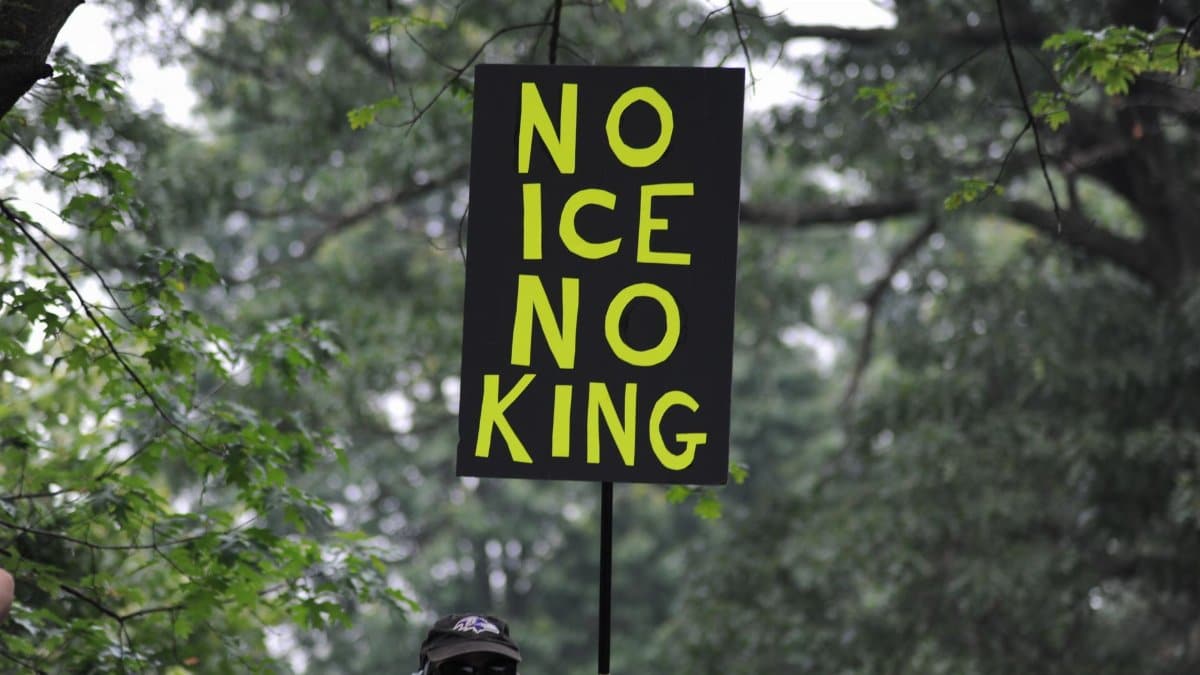 A protest sign with bold letters seen during a demonstration in Baltimore, surrounded by dense tree foliage.