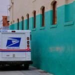A USPS delivery truck parked in a vibrant urban alley beside colorful architecture.
