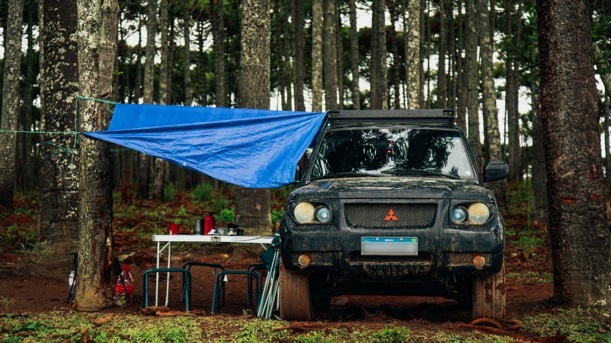 A rugged off-road vehicle parked in a forest clearing, set up for a camp with tarp and table.