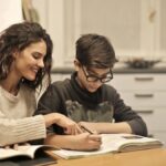 Cheerful young woman helping younger brother with homework while sitting together at table with books at home