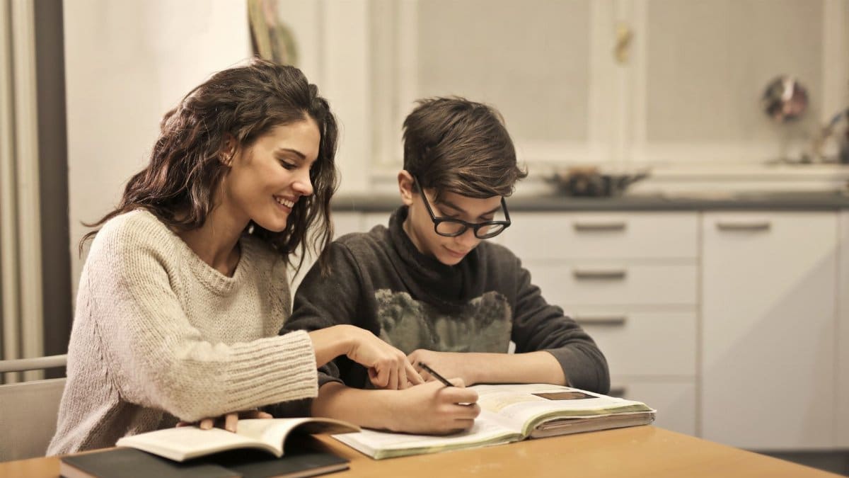 Cheerful young woman helping younger brother with homework while sitting together at table with books at home