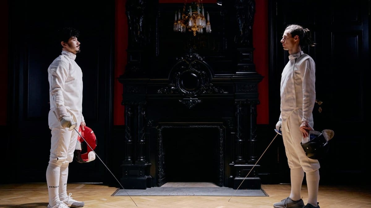 Two male fencers face off in an ornate room, showcasing a poised and disciplined sword duel indoors.