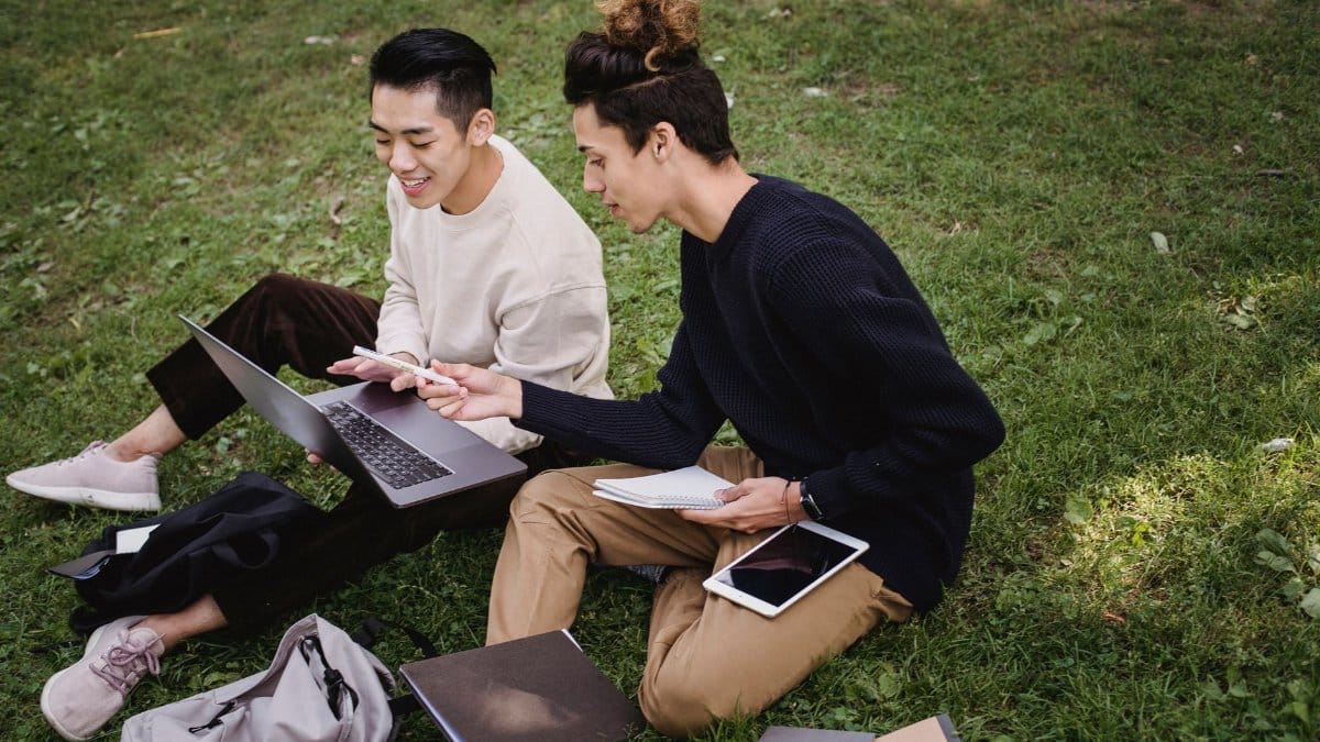 High angle full length ethnic male students in casual clothes browsing laptop and discussing college project while sitting together on grassy meadow in park