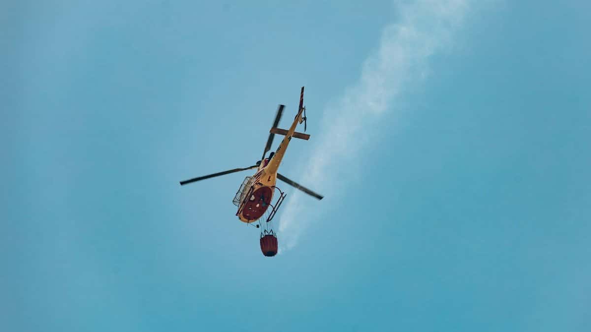 Helicopter performing aerial firefighting with water bucket against a clear sky in Braga, Portugal.