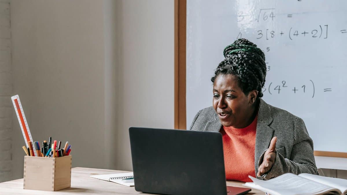 African American female teacher talking during class on netbook while sitting at desk with books and stationery in classroom