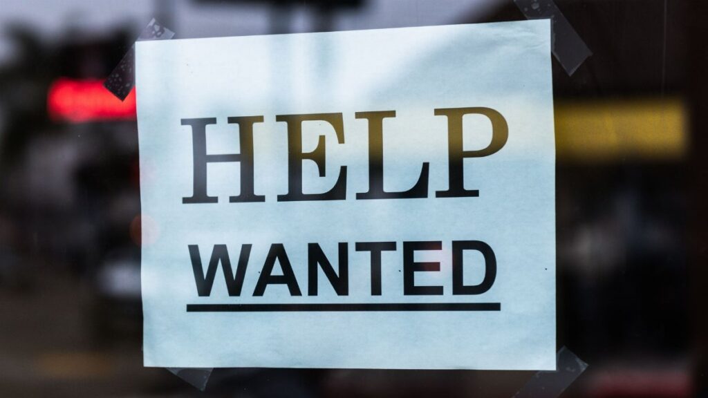 Close-up of a Help Wanted sign taped to a glass window in a storefront.