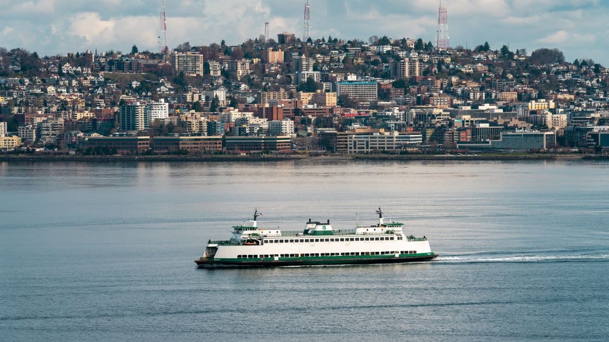 A Washington State Ferry sails on the calm water with Seattle skyline in the background.