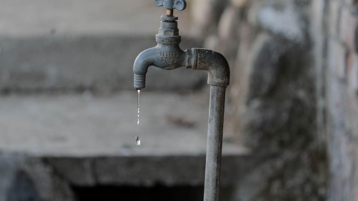 A close-up of a rustic outdoor faucet dripping water against a natural, textured background, symbolizing scarcity.