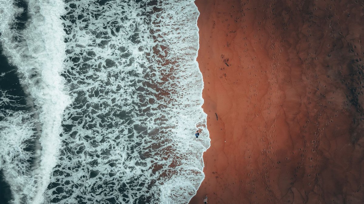 Aerial view of distant unrecognizable tourist standing in foamy water of powerful wavy ocean near sandy beach