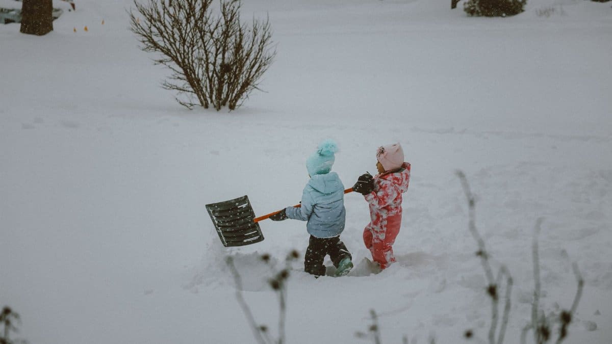 Two children in winter outfits play and shovel snow in a snowy landscape, enjoying winter fun.