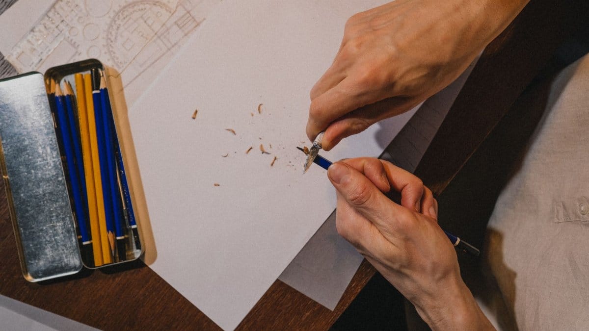 A close-up shot of a person sharpening a pencil over paper, surrounded by drawing tools indoors.