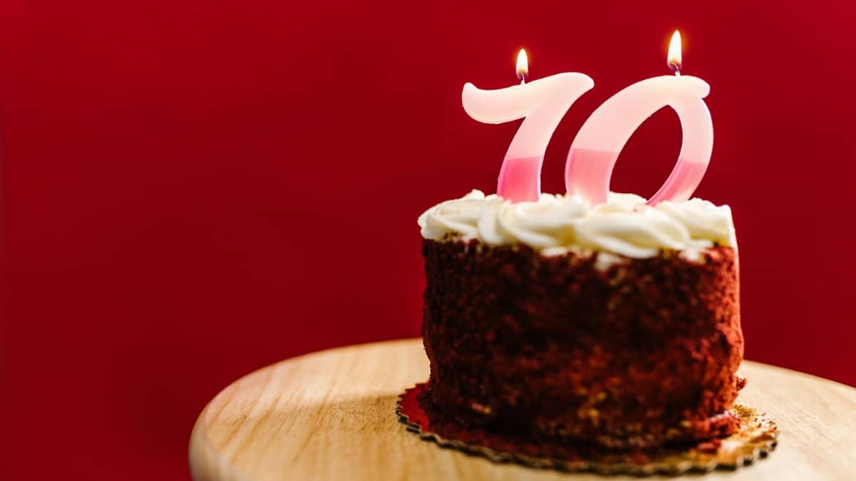 Delicious red velvet cake with number 70 candles on a wooden stand against a red backdrop.