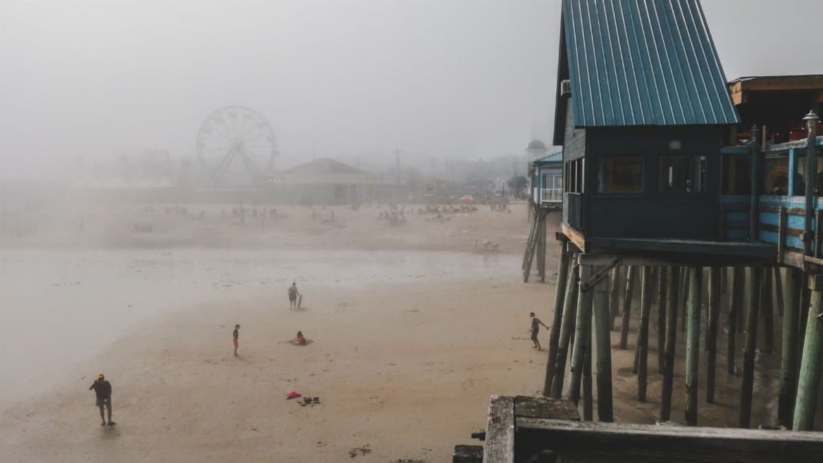 A foggy day on a beach with a Ferris wheel and pier extending over the sand. People scattered across the shore.