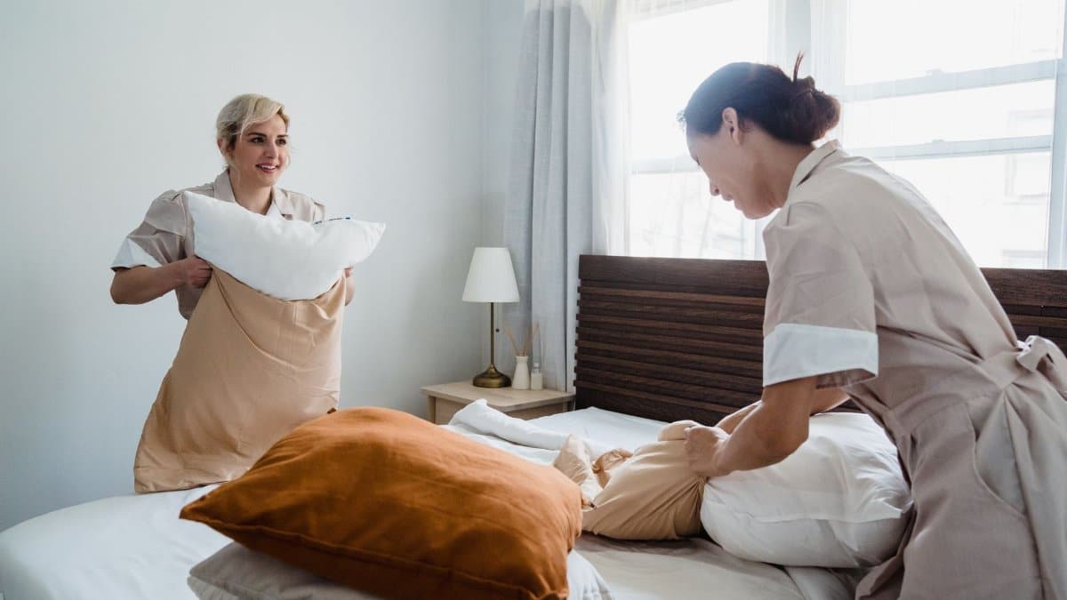 Two women in uniforms arranging bed linens in a clean and bright bedroom.