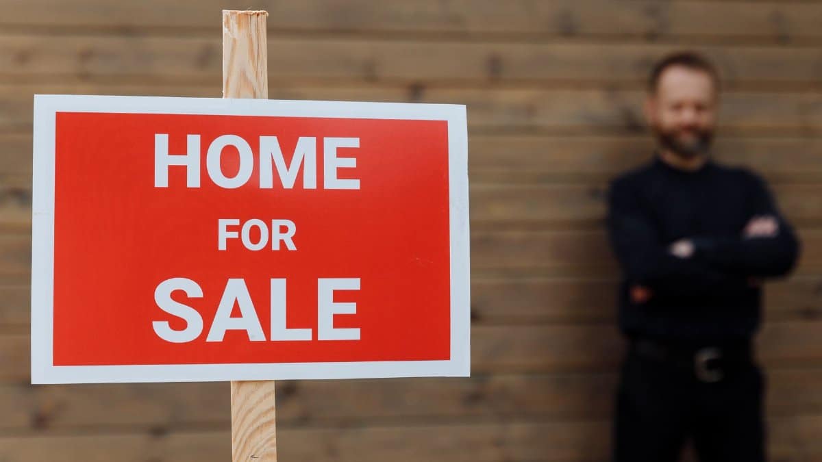 A confident real estate agent stands by a home for sale sign against a wooden wall.