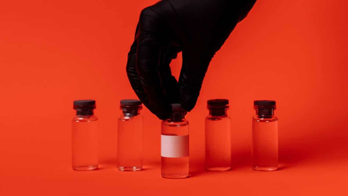 Person wearing black glove grabbing a vaccine vial against a bright red backdrop.