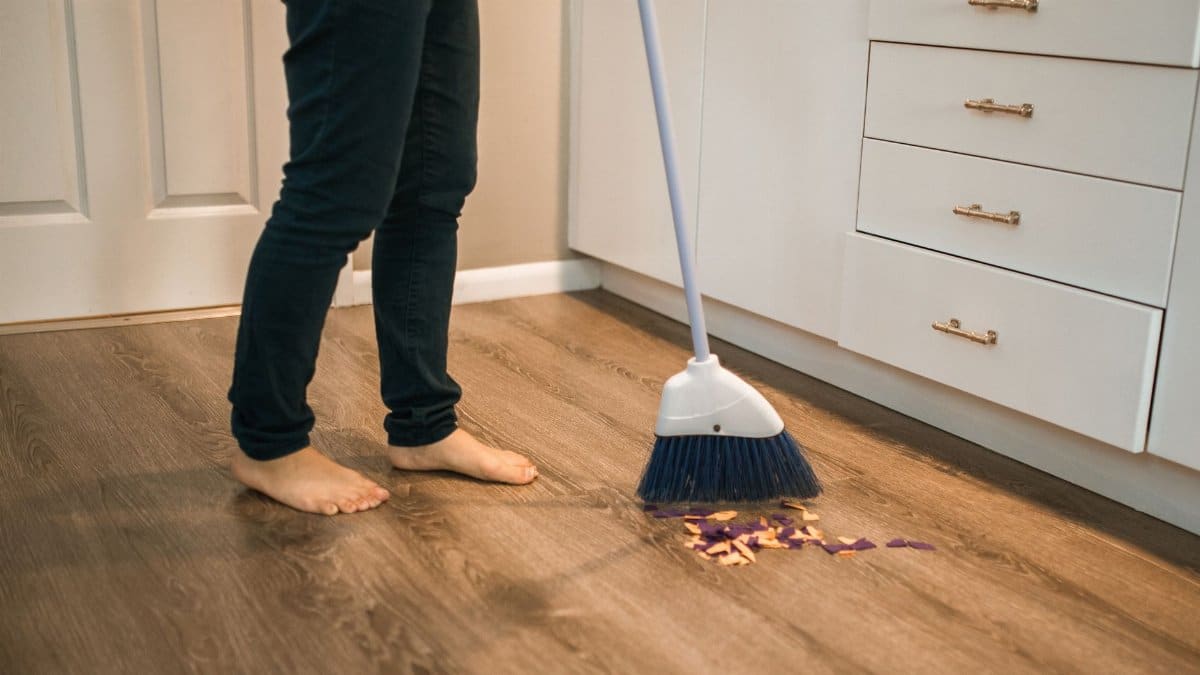 Person sweeping wooden kitchen floor with a broom, showing daily cleaning routine.