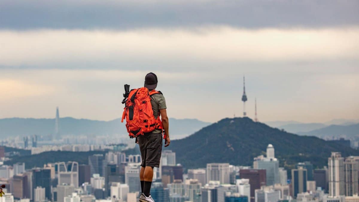Man with backpack observing Seoul cityscape from mountain peak.