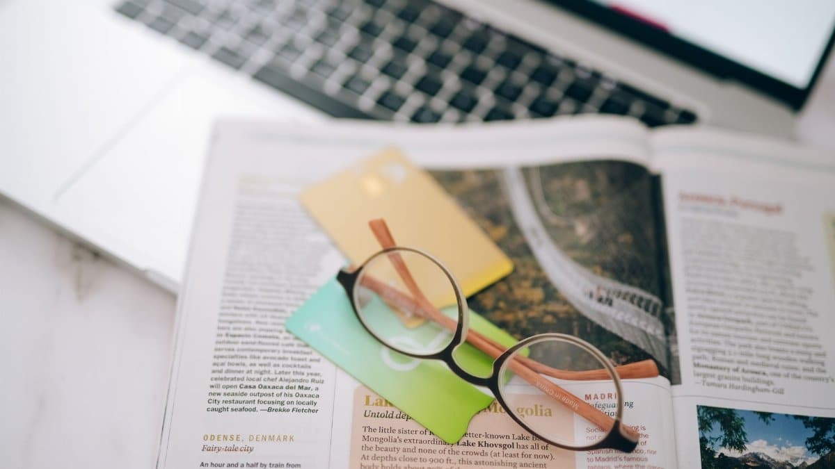 Overhead view of reading glasses, credit cards, and a magazine with a blurred laptop in the background.