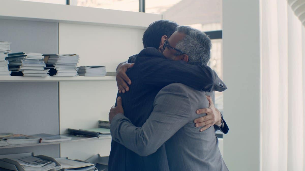 Two businessmen in suits share a warm hug in a sunlit office.